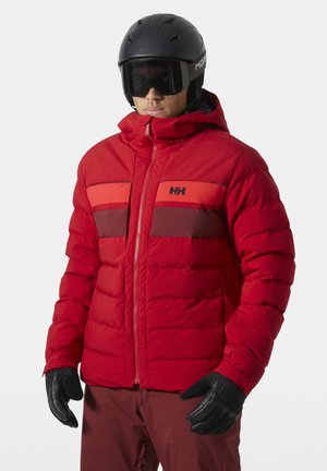 Persona vistiendo una chaqueta de invierno acolchada roja con rayas negras y rojo oscuro, casco de esquí negro, gafas y guantes negros.