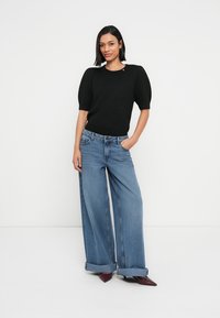 Pulover negru tricotat, cu mâneci scurte, asortat cu blugi albaștri largi din denim. Blugii au tiv pliat și o textură ușor uzată.