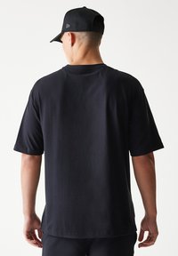 Μαύρο βαμβακερό T-shirt με άνετη εφαρμογή, κοντά μανίκια και στρογγυλή λαιμόκοψη, εμφανιζόμενο από πίσω. Το καπέλο έχει επίπεδο γείσο και λογότυπο.