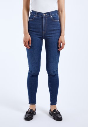 Vaqueros de mezclilla azul de cintura alta con un diseño ajustado, cinco bolsillos y detalles de costura en contraste. Llevados con zapatos mocasines negros.