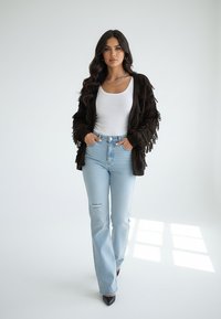 Brązowy kardigan z frędzlami nałożony na biały top na ramiączkach, w połączeniu z jasno niebieskimi jeansami z szerokimi nogawkami z rozdarciem na kolanie oraz czarnymi szpiczastymi szpilkami.