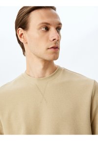 Helle beige Langarmshirt aus strukturierter, gerippter Stoffqualität. Mit Rundhalsausschnitt und einem dekorativen Kreuzstichdetail auf der Brust.