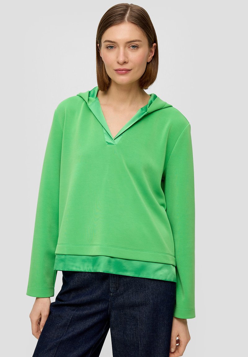 Groene hoodie van zacht materiaal, met een V-hals, lange mouwen en een ontspannen pasvorm, gedragen over donkerblauwe jeans.