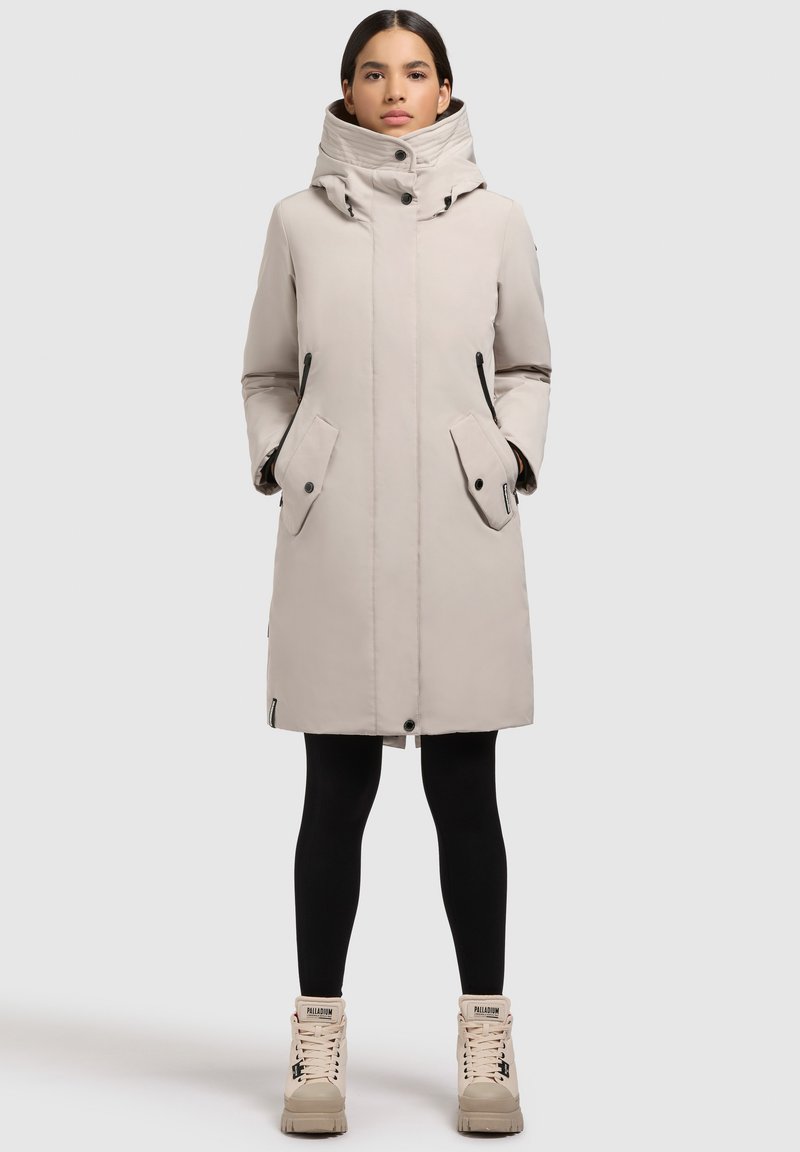 Vrouw staat met handen in de zakken, draagt een beige winterjas, zwarte leggings en beige hoge sneakers tegen een effen achtergrond.