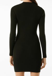 Schwarzes, geripptes Strickkleid mit langen Ärmeln, hohem Halsausschnitt und figurbetonter Silhouette. Der Stoff weist ein strukturiertes Muster auf.
