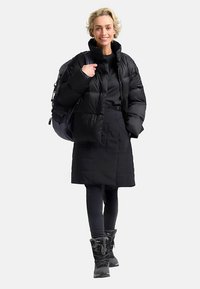 Zwarte donsjas met een hoge kraag, lange mouwen en een doorgestikt ontwerp. Gecombineerd met zwarte leggings en geïsoleerde laarzen. Draagt een rugzak.