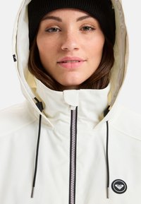 Witte waterdichte jas met een capuchon, voorzien van een rits aan de voorkant met roze accenten, zwarte koordjes en een ronde logo aan de linkerkant.