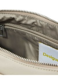 Borsa in tessuto beige con apertura a zip, caratterizzata da un interno liscio, un piccolo scomparto con zip e un'etichetta bianca con testo giallo.