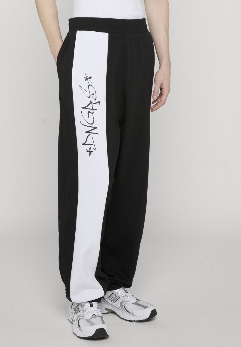 Person wearing black sweatpants with a white vertical stripe and graffiti-style "DNGRS" text on the side, paired with white sneakers.