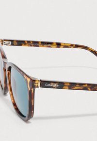 Occhiali da sole tartarugati con lenti sfumate blu. Design con montatura quadrata. Presentano il logo Calvin Klein sulla stanghetta.