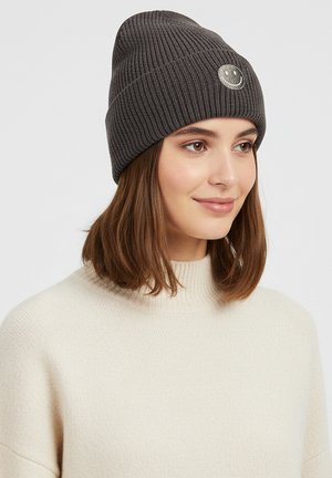 Gray ribbed knit beanie with a round silver smiley face pin detail, worn with a cream-colored sweater. Soft texture, fitted design.