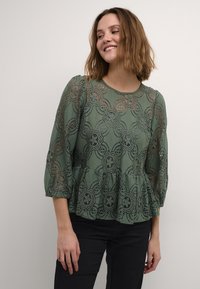 Zelený peplum top z krajky s květinovými vzory, s rukávy 3/4 a kulatým výstřihem. Spárováno s černými kalhotami, které zvýrazňují texturovaný design.