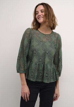 Zelený peplum top z krajky s květinovými vzory, s rukávy 3/4 a kulatým výstřihem. Spárováno s černými kalhotami, které zvýrazňují texturovaný design.