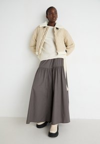 Beige quilted bomber jacket with a fur collar, cream sweater, and dark gray pleated skirt. Model wears beige chunky-sole boots.
