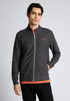 Uomo che indossa una giacca grigia scura con zip sopra una maglietta arancione, in piedi con una mano in tasca contro uno sfondo chiaro e uniforme.