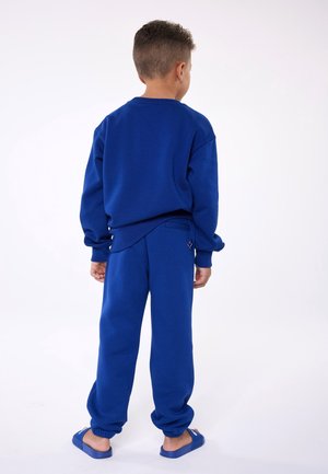 Kind staat met de rug naar de kijker, draagt een blauwe trui, blauwe joggingbroek en bijpassende slide sandalen, op een effen witte achtergrond.