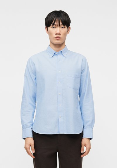 Jeune homme portant une chemise bleu clair à boutons avec des manches longues et un pantalon noir, debout devant un fond blanc uni.