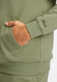 Sweat à capuche vert olive en tissu doux, avec une poche avant et des poignets côtelés. Logo sur la manche en fil contrastant.