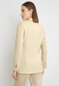Cream-colored tailored blazer with a structured fit, notched lapels, and golden button accents on the sleeves. Rear view showcases clean lines.