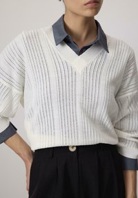 Pull en tricot blanc avec des côtes verticales et un col en V, associé à une chemise bleu à col en dessous, complété par un pantalon noir.