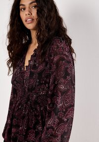 Femme avec de longs cheveux bouclés et foncés, portant une robe noire avec des motifs paisley et floraux roses, se tenant devant un fond uni clair.