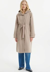 Beige leichter Trenchcoat mit Kapuze, tailliertem Gürtel und seitlichen Taschen. Hat eine glatte Textur und gepolsterte Ärmelabschlüsse.