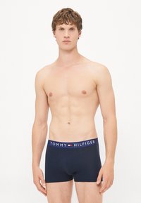 Marineblaue Boxershorts mit einfarbigem Design, ausgestattet mit einem weißen Bund, der mit der Aufschrift "TOMMY HILFIGER" in fettgedruckten Buchstaben versehen ist.
