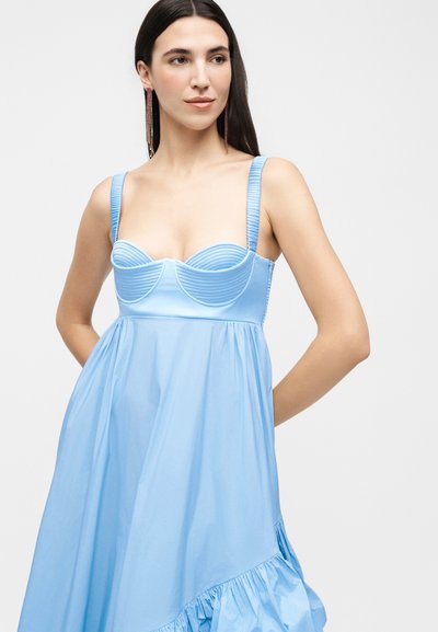 Γυναίκα που φοράει αμάνικο ανοιχτό μπλε φόρεμα με δομημένο μπούστο, φαρδιές τιράντες και βολάν στο τελείωμα, πόζαρε με τα χέρια πίσω από την πλάτη.