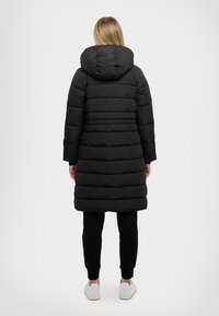 Cappotto imbottito nero con cappuccio, caratterizzato da sezioni trapuntate orizzontali e un retro leggermente più lungo. Indossato con pantaloni neri e sneaker bianche.