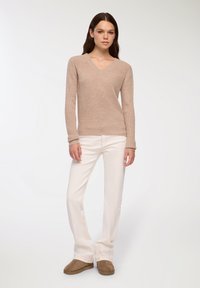 Beżowy sweter dzianinowy z dekoltem w serek, długimi rękawami i prążkowanymi mankietami, zestawiony z białymi dżinsami w stylu dzwonów i brązowymi zamszowymi mokasynami.