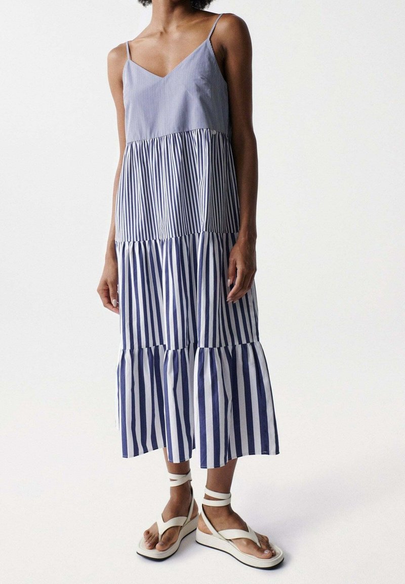 Pruhované bezramínkové šaty s rozšířenou spodní částí, s modro-bílými svislými pruhy. Nosené s bílými platformovými sandály.