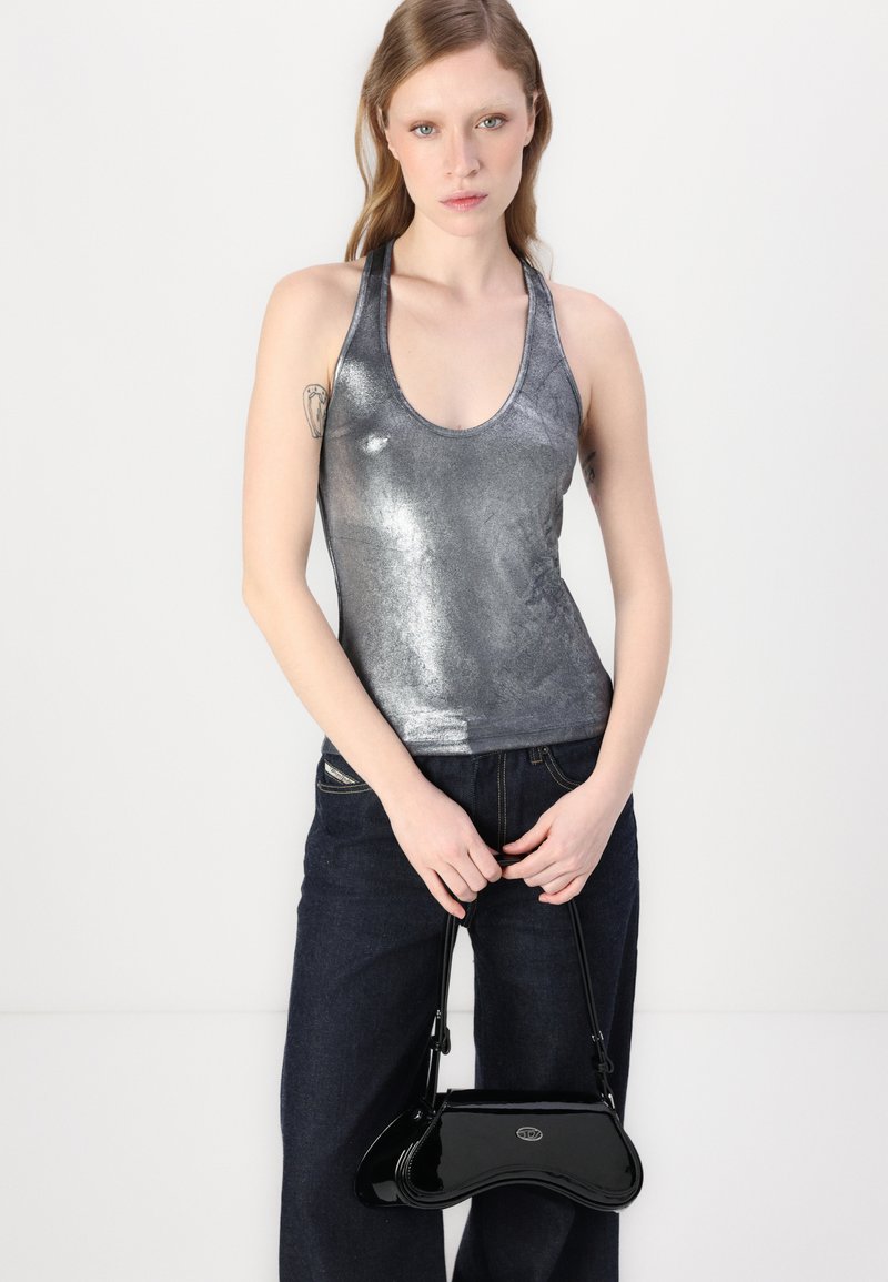 Metallisch silbernes Tanktop mit runder Ausschnitt, kombiniert mit dunklen Jeans. Das Modell trägt eine kleine schwarze Handtasche aus Lackleder.