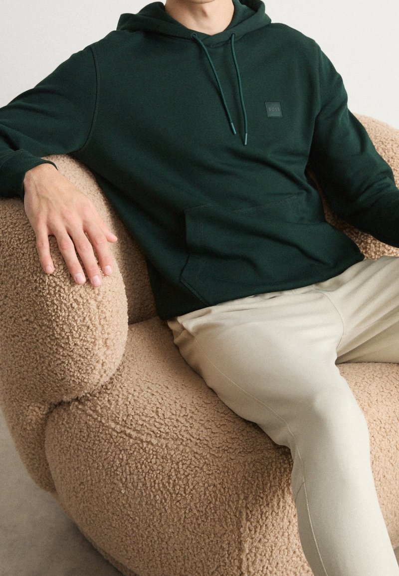 Homme portant un sweat à capuche vert foncé et un pantalon beige assis dans un fauteuil beige texturé, les bras reposant sur les accoudoirs.