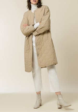 Femme portant un long manteau matelassé beige sur un col roulé blanc, un pantalon blanc court et des bottines beiges, debout les bras croisés.