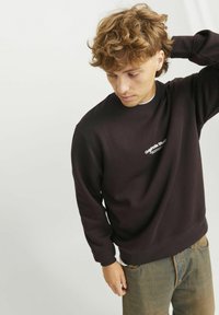 Zwarte sweatshirt van zachte stof met een ronde halslijn en witte tekst "Originals Studio". Het model draagt een verstoorde lichte jeans.
