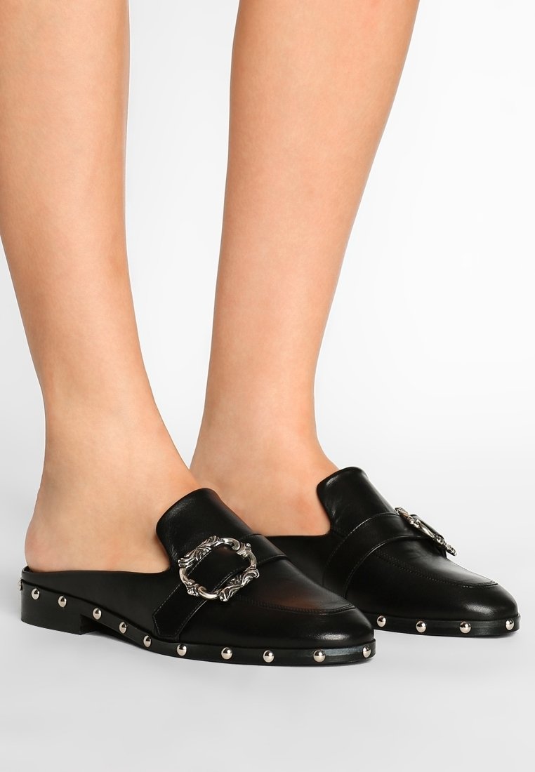 Mules en cuir noir à dos ouvert avec une accent de boucle et des clous argentés le long de la semelle. Texture lisse avec une forme de pied arrondi.