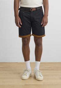 Mustad lühikesed püksid pruuni rullitud äärega, millel on kuldse pandlaga vöö, valgete sokkide ja heledate kingadega, seisab puidust põrandal.