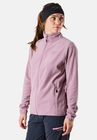 Fleece-takki vaaleanpunaisena, jossa on korkea kaulus, kaksi vetoketjullista taskua ja ribbireunukset. Rintamuksessa on hillitty brändilogo.