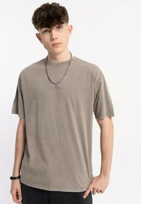 Młody mężczyzna z krótkimi, kręconymi włosami, ubrany w przetartą, brązową, oversize'ową koszulkę, naszyjnik z łańcuchem i czarne spodnie, stojący na tle białym.