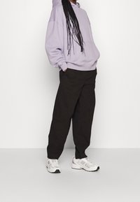 Fioletowy, oversize'owy hoodie z kieszenią kangurką, połączony z czarnymi spodniami o szerokiej nogawce i białymi sportowymi trampkami. Gładka tekstura materiału.