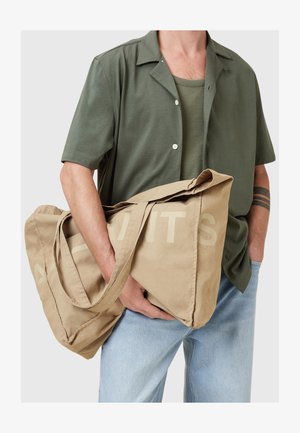 Tan lerret tote bag med store hender, med blokkbokstaver trykt på siden. Bagen har en myk tekstur og romslig design.