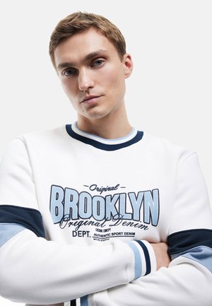 Biała bluza z grafiką "Brooklyn" w kolorze niebieskim, z prążkowanymi, granatowymi mankietami i okrągłym dekoltem. Materiał bawełniany, luźny krój, minimalistyczny design.
