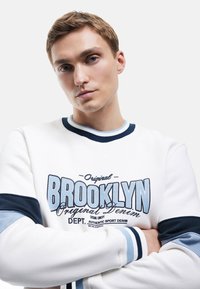 Sudadera blanca con gráfico de "Brooklyn" en azul, puños de canalé en navy, y escote redondo. Material de algodón, corte casual, diseño minimalista.