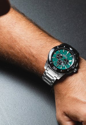 Srebrna ura iz nerjavečega jekla z zeleno teksturirano številčnico in črnim obročem. Chronograph podmerniki in masivni kazalci na 2, 4, 8.