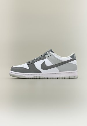 Niski sneaker Nike w kolorze białym, jasno szarym i ciemno szarym z szarym logo, sznurówkami i białą podeszwą na beżowym tle.