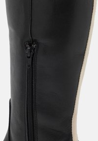 Boot en cuir noir avec une fermeture éclair sur le côté et un panneau beige contrastant. Texture lisse avec des coutures détaillées le long des joints.
