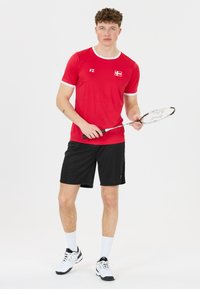 Nuori mies punaisessa urheilupaidassa ja mustissa sortseissa pitää badmintonmailaa, seisten valkoista taustaa vasten.