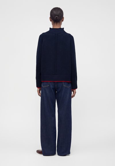 Personne avec les cheveux en chignon portant un pull bleu marine foncé avec des poignets rouges et un jean large bleu foncé, debout de dos sur un fond uni.