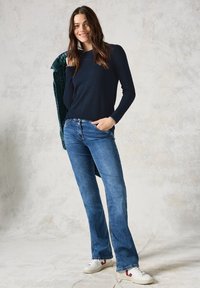 Maglietta a maniche lunghe blu navy, jeans a zampa d'elefante con cucitura sottile e sneaker bianche con dettagli rossi. Modella in posa contro uno sfondo chiaro e testurizzato.