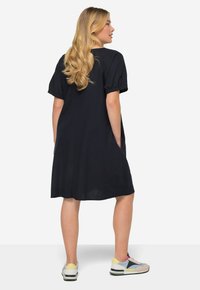 Tmavě modré šaty s krátkým rukávem a volným střihem, vybavené bočními kapsami a délkou pod kolena, doplněné vícebarevnými teniskami.